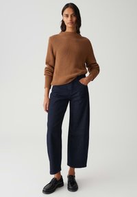 Frau trägt einen braunen gerippten Pullover, dunkle weit geschnittene Jeans und schwarze Schnürschuhe, steht mit einer Hand in der Tasche vor einem schlichten Hintergrund.