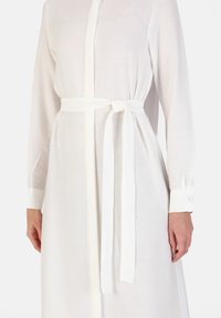 White, long-sleeve dress in a light, textured fabric with a belted waist. Features buttoned cuffs and a slightly open collar.