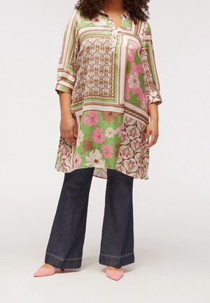 Femme portant une tunique à motifs floraux et géométriques avec des manches trois-quarts, un jean large foncé et des escarpins roses à bouts pointus.