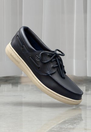 Navy-blaue Lederbootsschuhe mit glatter Oberfläche, beiger Gummisohle und tonal abgestimmten Schnürsenkeln. Mit geprägtem Logo an der Seite.