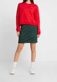 Röd crewneck sweatshirt med logotyp, parat med en grön mockaskirt med frontdragkedja. Vita sneakers och strumpor fulländar looken.