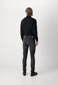 Suéter negro de cuello alto ajustado para hombre combinado con jeans gris oscuro que presentan un logo de CK en la cintura trasera, y zapatos negros.