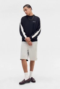 Muž s vrkočmi, oblečený v čiernej mikine s bielymi pruhmi, bielych sieťovinových šortkách, bielych ponožkách a hnedej mokasínovej obuvi, stojaci na bielom pozadí.