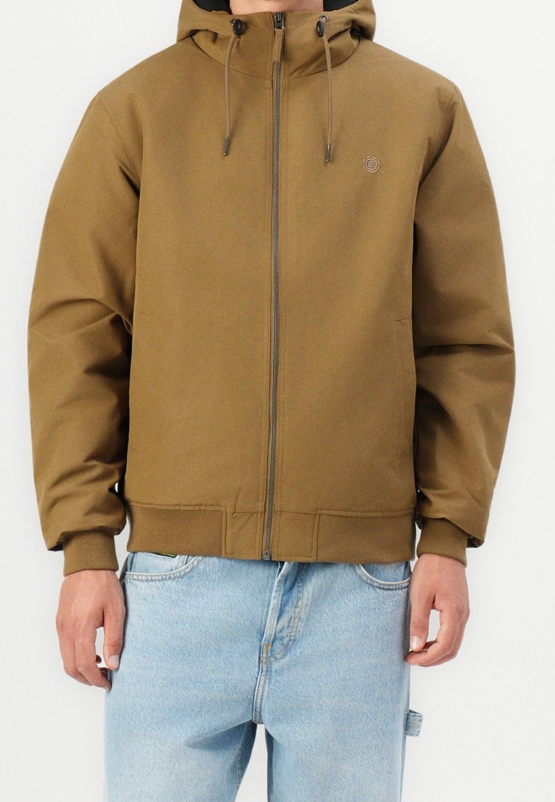Homme portant une veste à capuche marron avec fermeture éclair et cordons, associée à un jean bleu clair, debout devant un fond uni.