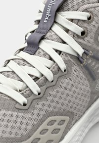 Světle šedá sportovní bota s síťovinovým svrškem, bílými tkaničkami, texturovaným vzorem a flexibilní podrážkou, která má šedou logovou nášivku nad tkaničkami.