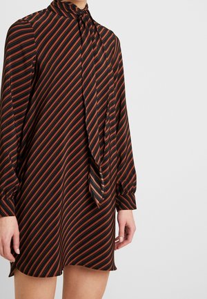 Frau trägt ein schwarzes Kleid mit diagonalen rost-orangefarbenen Streifen, langen Ärmeln, mit Bündchen an den Handgelenken und einer verbundenen Krawatte, die vorne herunterhängt.