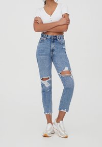 Jeans z wysokim stanem w niebieskim kolorze, z przetarciami, zwężanymi nogawkami i surowym wykończeniem. Połączone z białymi trampkami i białą bluzką z krótkim rękawem.