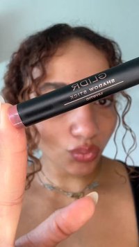Jeune femme tenant un crayon fard à paupières noir Glidr Shadow Stick Mystic près de la caméra, avec des cheveux bouclés et portant un collier.