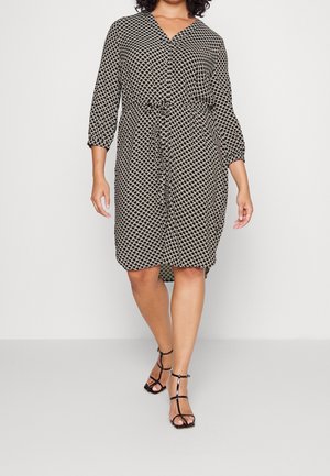 Kobieta ubrana w czarno-białą sukienkę do kolan w wzory z rękawami za trzy czwarte i czarne, paskowane sandały na wysokim obcasie, idzie do przodu.