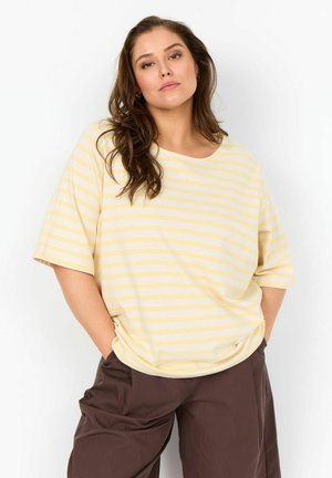 Frau mit langen braunen Haaren, die ein locker sitzendes gelb-weiß gestreiftes T-Shirt und braune Hose trägt, steht mit den Händen in den Taschen vor weißem Hintergrund.