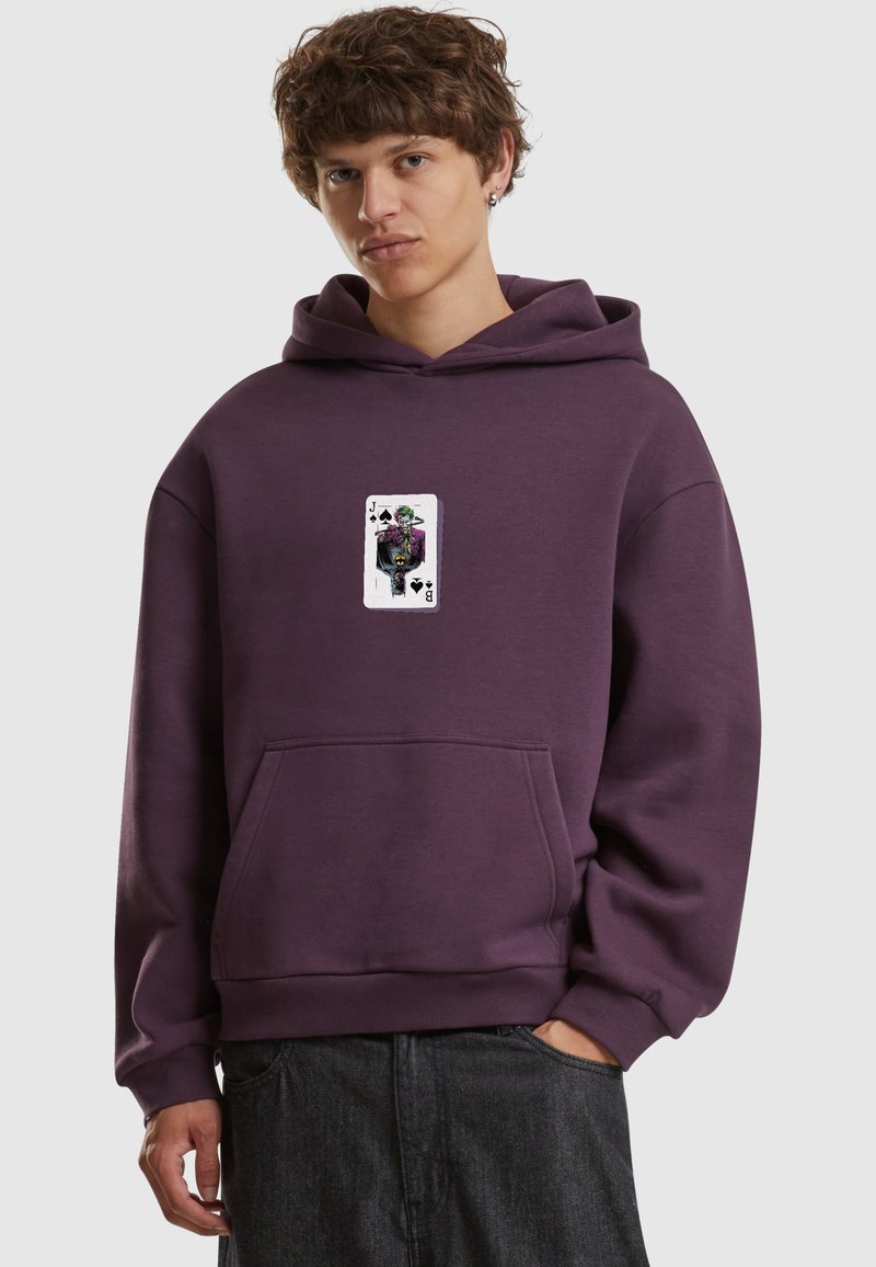 Jeune homme portant un sweat à capuche violet avec un motif du valet de pique sur la poitrine, debout avec une main dans la poche.