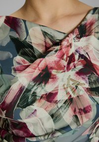 Blumenstoff mit großen, abstrakten rosa und weißen Blüten sowie grünen Blättern. Weiche Textur und hängendes Design auf einem sanften blauen Hintergrund.