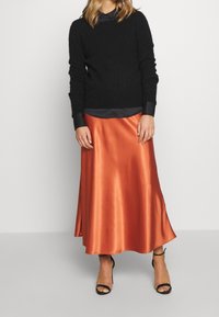 Svart stickad tröja, grå skjorta med krage och en lång kjol i orange satin. Svarta sandaler med remmar fulländar outfiten. Slät textur med lätt lyster.