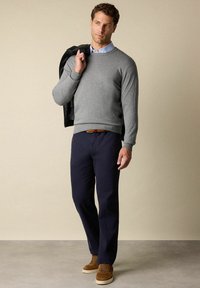 Grijze pullover met geribbelde manchetten en zoom, marineblauwe broek en bruine suède schoenen. Zwarte jas over de schouder gedrapeerd, lichte achtergrond.