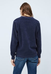 Femme aux cheveux bruns portant un sweat-shirt bleu marine et un jean bleu, vue de dos sur un fond blanc uni.