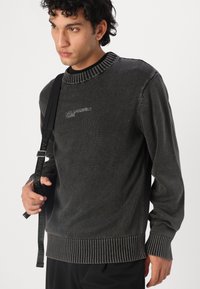 Mörkgrå tröja med ribbad krage och nederkant, texturerad tyg och subtil märkning på bröstet. Modellen håller i ett rem.