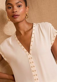 Blusa beige con scollo a V e maniche corte, dotata di accenti in metallo tono oro lungo la parte anteriore. Tessuto liscio e leggero.