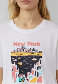 T-shirt en coton blanc avec un graphique coloré d'une silhouette de ville et des bateaux sur l'eau, avec "New York" imprimé en rose de manière visible.