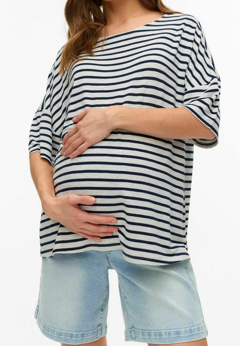 Randig oversized topp i grått och marinblått, kombinerad med ljusblå denimshorts. Toppen har vida ärmar, och modellen håller varsamt om en gravid mage.