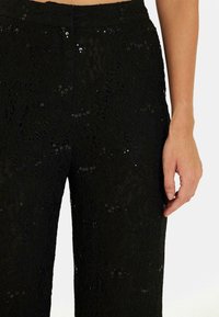 Zwarte kant broeken met een gestructureerd bloemenpatroon en pailletten, met een hoge tailleband en een wijde pijpdesign. Hand geplaatst nabij de taille.