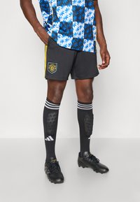 Sorte fodboldshorts med gule detaljer, mønstrede blå og sorte trøje, sorte sokker med hvide striber og sorte fodboldstøvler.