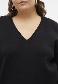 Kobieta w czarnym swetrze z dekoltem w szpic i złotych skręconych kolczykach w kształcie kół, z falującymi brązowymi włosami.