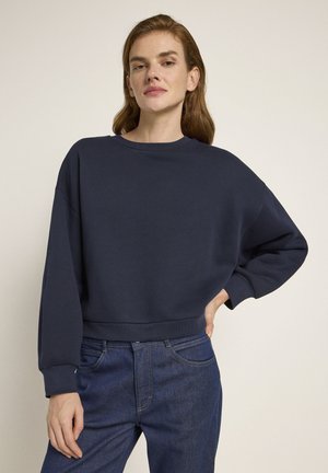 Navy blue cropped sweatshirt with a crew neckline and dropped shoulders. Soft texture, ribbed cuffs, and a relaxed fit, paired with blue jeans.