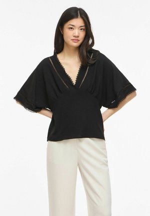 Femme aux longs cheveux foncés portant un chemisier noir à col en V avec bordure en dentelle et manches larges, associé à un pantalon beige clair, debout les mains sur les hanches.