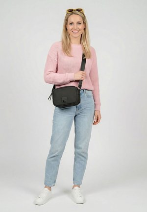 Lachende vrouw in een roze trui, lichtblauwe spijkerbroek, witte sneakers, met een zwarte schoudertas en een zonnebril op haar hoofd, tegen een effen achtergrond.