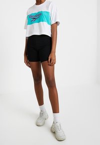 Bílý krátký trik s modro-zeleným a fialovým logem Reebok, doplněný černými šortkami s vysokým pasem a světle šedými teniskami.