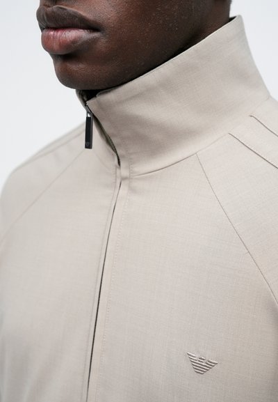 Beige ζακέτα με φερμουάρ και ψηλό γιακά, με διακριτές ραφές και ένα μικρό κεντημένο λογότυπο σε σχήμα φτερών στη θηλή.