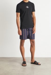 Svart bomull t-shirt med logotyp på bröstet, marinblå randiga badbyxor och svarta slidesandaler på en neutral bakgrund.