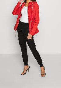 Veste à capuche rouge avec cordons, haut blanc ajusté, pantalon cargo noir et sandales à talons hauts noires à brides. Textures lisses et design décontracté.