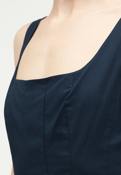 Κοντινό πλάνο ενός ατόμου που φοράει ένα σκούρο ναυτικό φόρεμα χωρίς μανίκια με τετράγωνη λαιμόκοψη και ορατές ραφές.