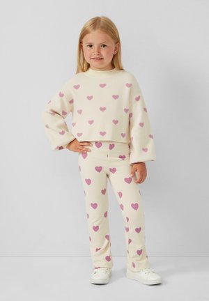 Cremfarbener, gekürzter Pullover und passende Hose, beide mit rosa Herzmustern. Weicher Stoff, lockere Passform und weiße Sneaker.