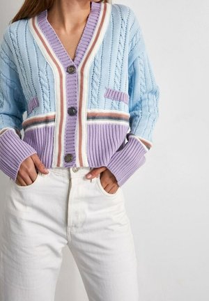 Femme portant un cardigan en maille bleu pastel et violet avec des boutons, une bordure rayée et un jean blanc taille haute, les mains dans les poches.