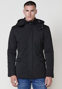 Cappotto nero con cappuccio, dotato di chiusura a bottoni, tasche sul petto doppie, colletto regolabile e tessuto liscio e texturizzato.