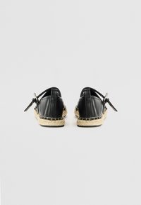 Zwarte leren espadrilles met een geweven jute zool, voorzien van een platte hak en een strikdetail aan de achterkant. Gladde textuur met minimale accenten.