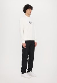 Hvid sweatshirt med sort logotekst, parret med mørk denimjeans og hvide sneakers. Modellen står foran en ensfarvet baggrund.