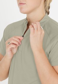 Ljusgrön zip-up-tröja med en texturerad tyg, ståkrage och en delvis dragkedja. Händerna visar silvriga ringar medan de justerar dragkedjan.