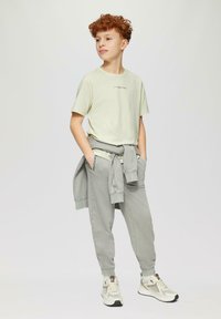 Lichtgroene t-shirt met kleine tekst, grijze sweatpants met boorden, grijze sweatshirt om de taille geknoopt, en beige sneakers met accenten.