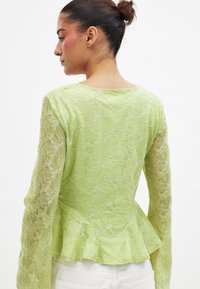 Heleda peplum-särk heledas rohelises värvitoonis lillemustriga. Omab pikki varrukaid ja laienevat hemmi, näidates delikaatset, läbilaskvat tekstuuri.