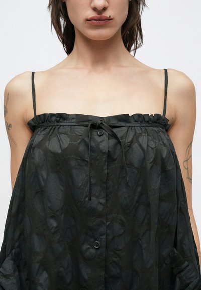 Μαύρο αμάνικο τοπ με διακριτικό υφασμένο σχέδιο, βολάν στη γραμμή του λαιμού, ρυθμιζόμενα κορδόνια και κλείσιμο με κουμπιά στο μπροστινό μέρος. Ελαφρύ ύφασμα.