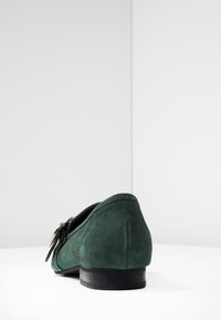 Chaussure slip-on en daim vert avec un petit talon, détail de nœud décoratif, texture lisse et bout arrondi. Design simple et élégant.