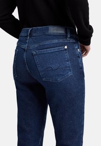 Jeans in denim blu scuro con una texture liscia, dotati di due tasche posteriori e un design a onda cucito sulla tasca sinistra.