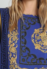 Frau in einem blauen Gewand mit filigranen goldenen und schwarzen barocken Blumen- und Scrollmustern, sichtbar von Hals bis zur Mitte der Brust.