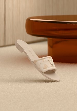 Sandale beige à bride en tissu avec un motif subtil, reposant sur un tapis crème près d'une table ronde couleur ambre.