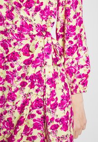 Robe jaune avec imprimé floral rose vif, manches longues et ceinture nouée à la taille, vue de la taille au poignet.