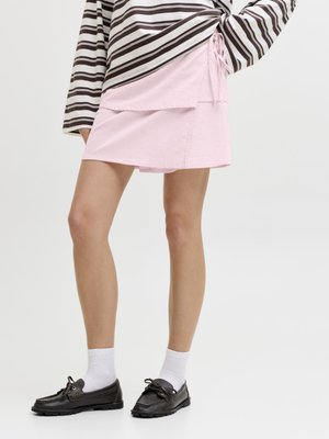 Kobieta ubrana w różową spódnicę na zakładkę, czarne mokasyny, białe skarpetki i długi rękaw w brązowo-białe paski, stojąca na jednolitym tle.