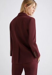 Donna bionda che gira la testa indossando una giacca su misura di un elegante color bordeaux scuro e pantaloni coordinati, su uno sfondo chiaro e neutro.